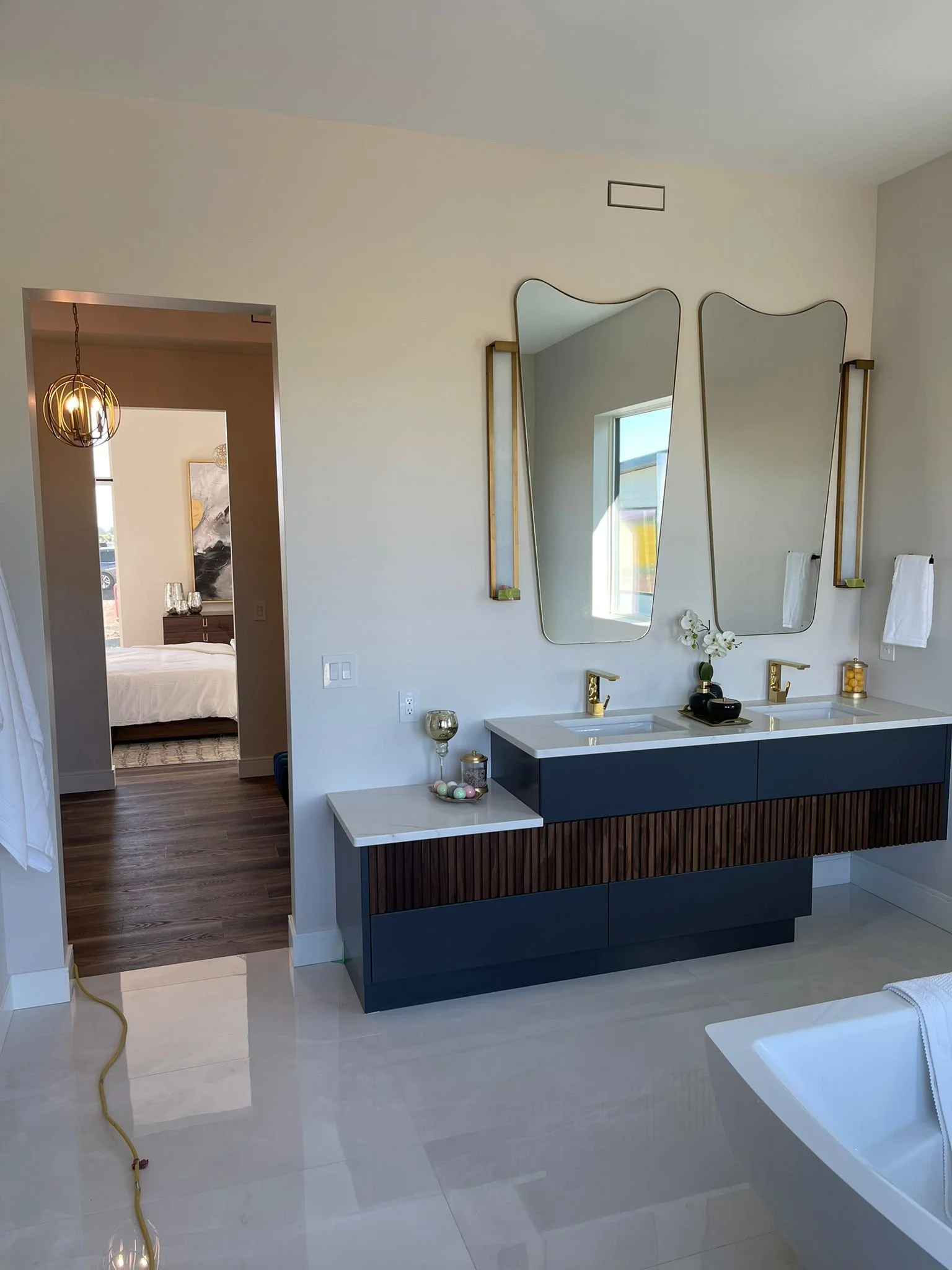 A modern bathroom with a dual sink vanity featuring gold fixtures, two irregularly shaped mirrors, a white orchid, and decorative items. There is a window with sunlight, and a view into a bedroom with a bed, artwork, and a hanging chandelier.
