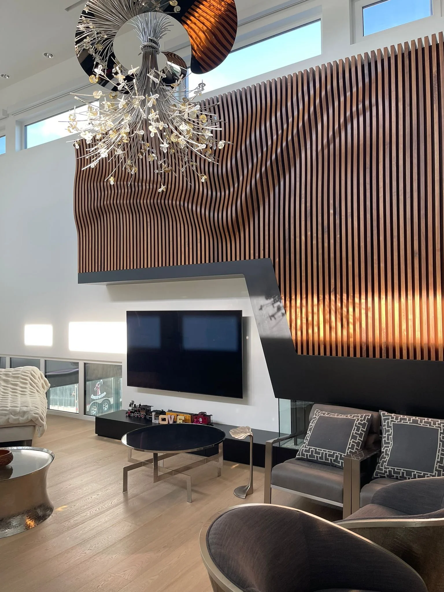 Modern living room with a large, decorative wooden slat wall, a contemporary chandelier, and a flat-screen TV mounted on the wall. There are chairs, a small round table, and a display with toy trains and the word 'LOVE' on a shelf.