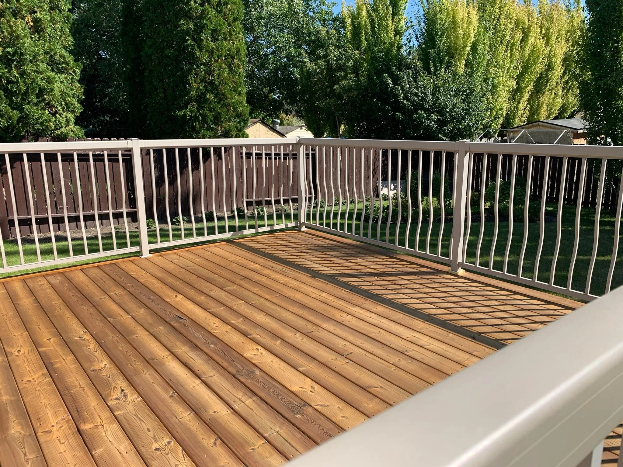 View of a backyard wooden deck with a white railing, green trees, and a clear blue sky.