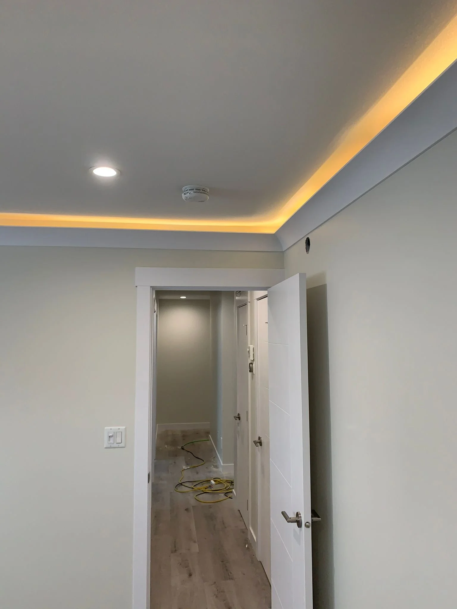 An interior view of a hallway with an open door leading to another room under construction. There is a smoke detector on the ceiling, electrical wires and tools on the floor, and a light switch on the wall.