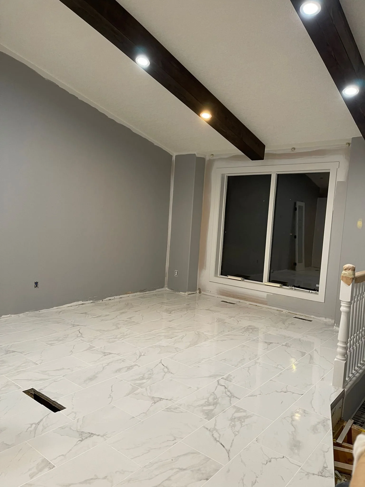 Empty room with white marble tile flooring, gray walls, a large window, and ceiling with dark wooden beams and recessed lighting.
