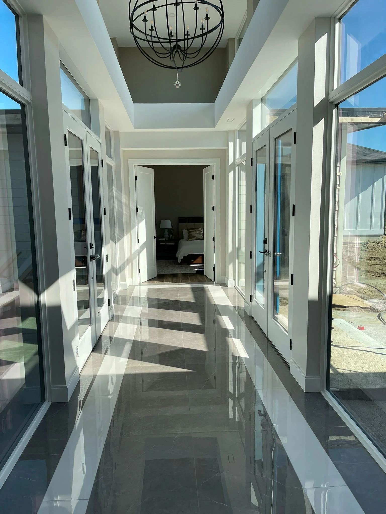 Bright sunlit enclosed porch with large glass windows on both sides, black chandelier hanging from ceiling, and doorway leading into a bedroom with visible bed and nightstand.