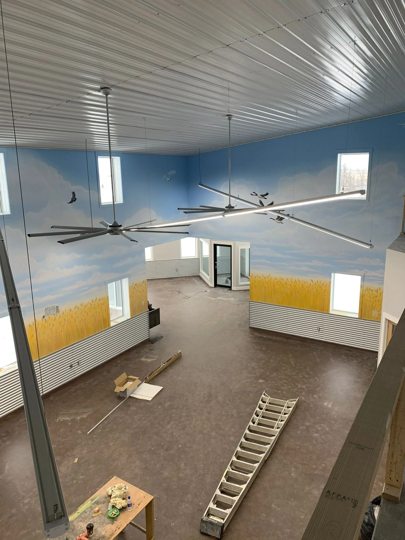 Interior of a partially constructed building with a mural of blue sky, white clouds, and flying birds painted on the walls. Ceiling has several modern ceiling fans, and the floor has construction materials and tools scattered around.