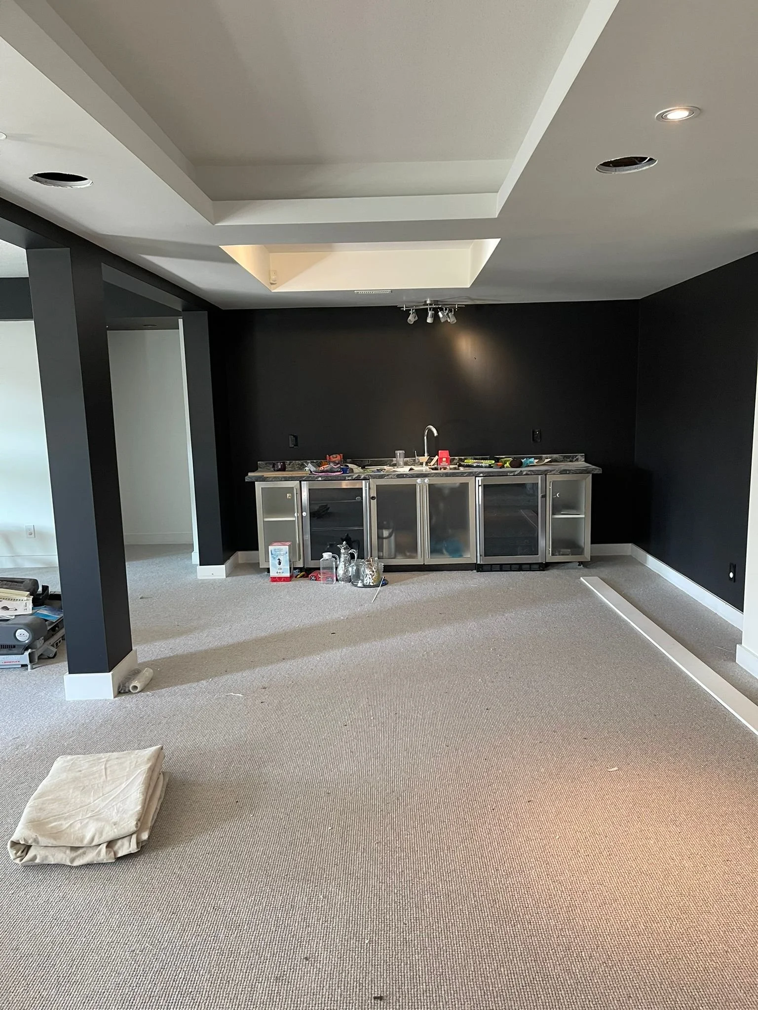 Unfinished kitchen with black walls, a ceiling with cutouts, a countertop, and an exposed cabinet frame. Tools and supplies are scattered on the counter, and a pillow or cushion is on the beige carpeted floor.