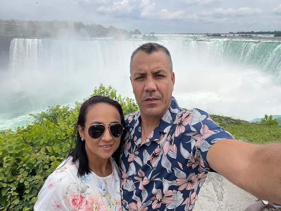 A couple takes a selfie in front of Niagara Falls, with lush green bushes and the waterfall in the background.
