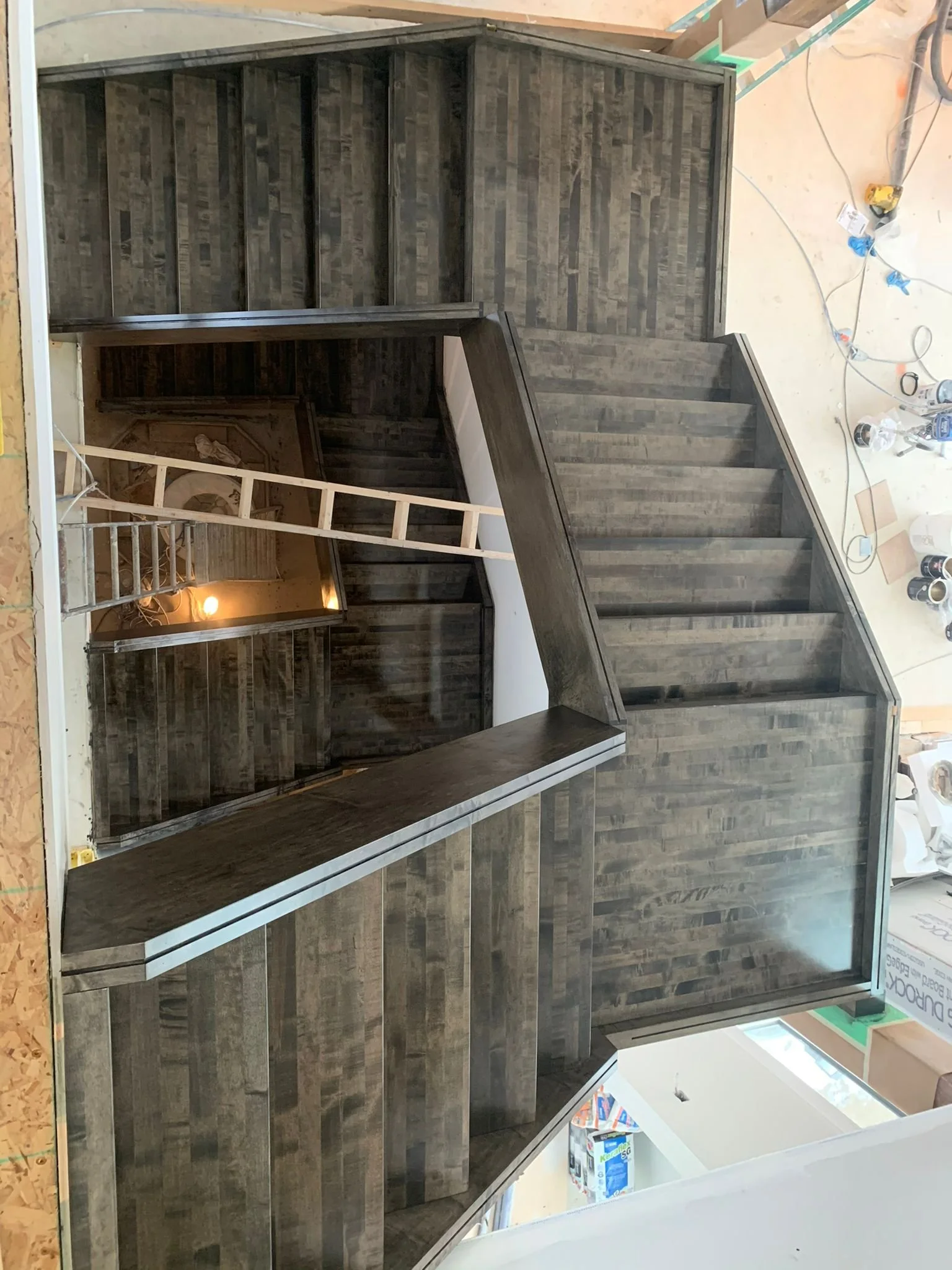 Construction site showing a partially built staircase with wooden steps and framing, and a wall covered in dark wood paneling. Tools and supplies are scattered on the floor around the construction area.