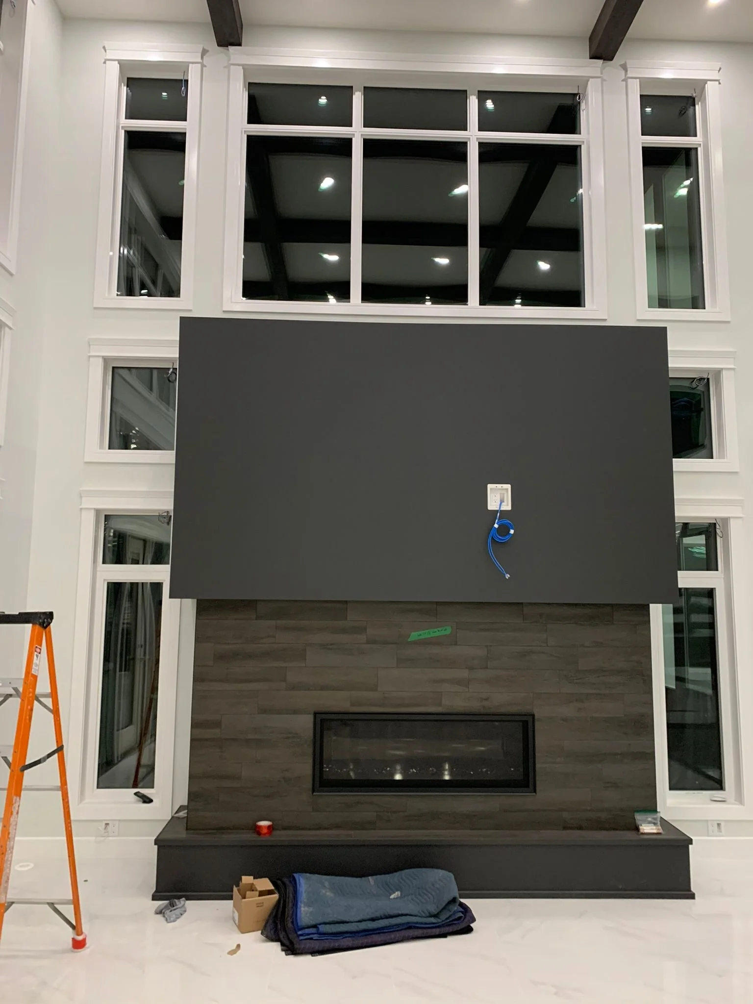 Living room with a large black fireplace, gray wooden paneling, and white-framed windows. Construction tools and materials are present, indicating ongoing home renovation or installation work.