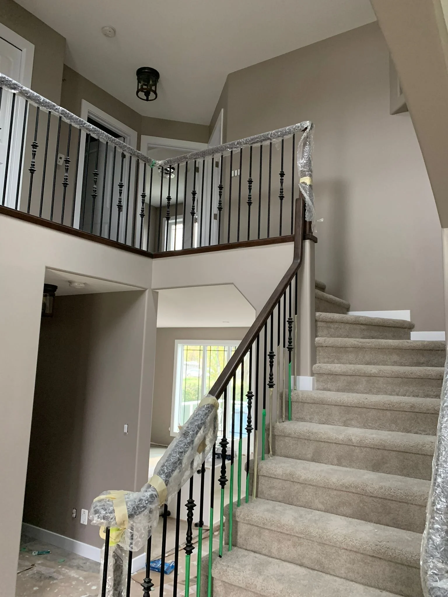 Interior view of a staircase under construction with new black metal railing, some parts wrapped in protective plastic, and a beige wall. There are nearby windows and some construction tools and materials on the floor.
