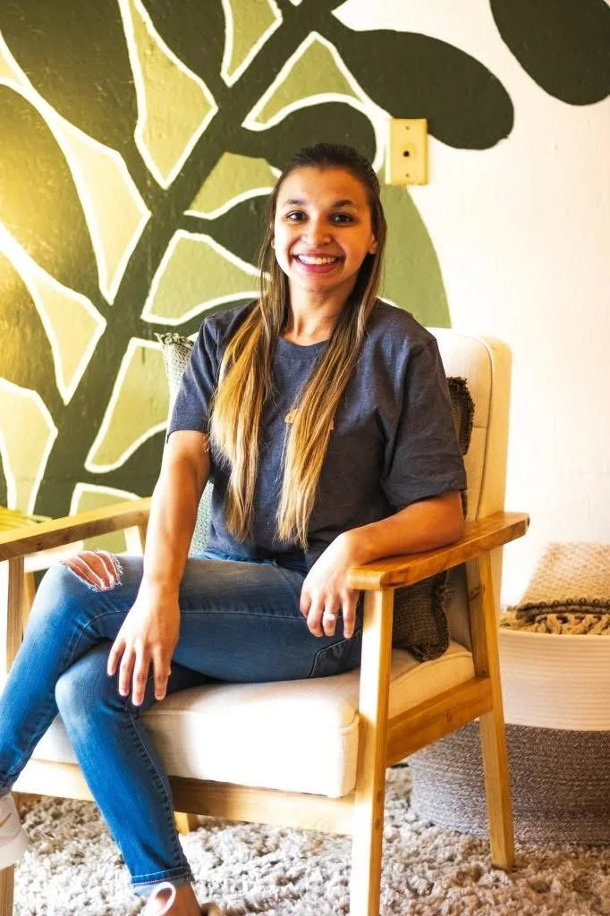 A young woman with long hair sitting on a wooden chair with cream-colored cushions, smiling and looking at the camera, in a room with a wall decorated with a large black and green abstract leaf pattern.
