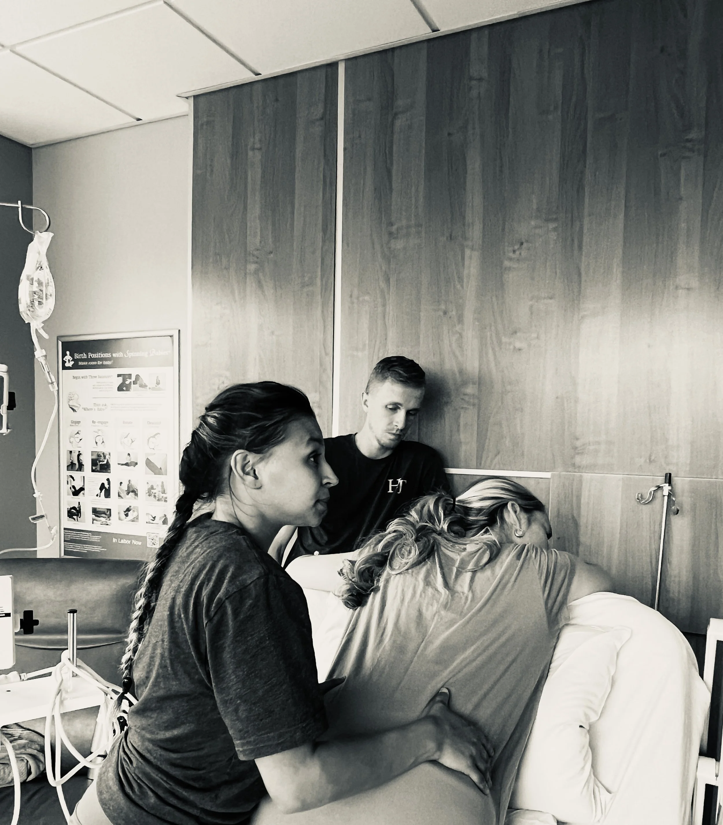 People in a hospital setting, with one person lying in a hospital bed, another standing next to the bed, all wearing dark clothing and medical masks, in a room with medical posters and equipment.