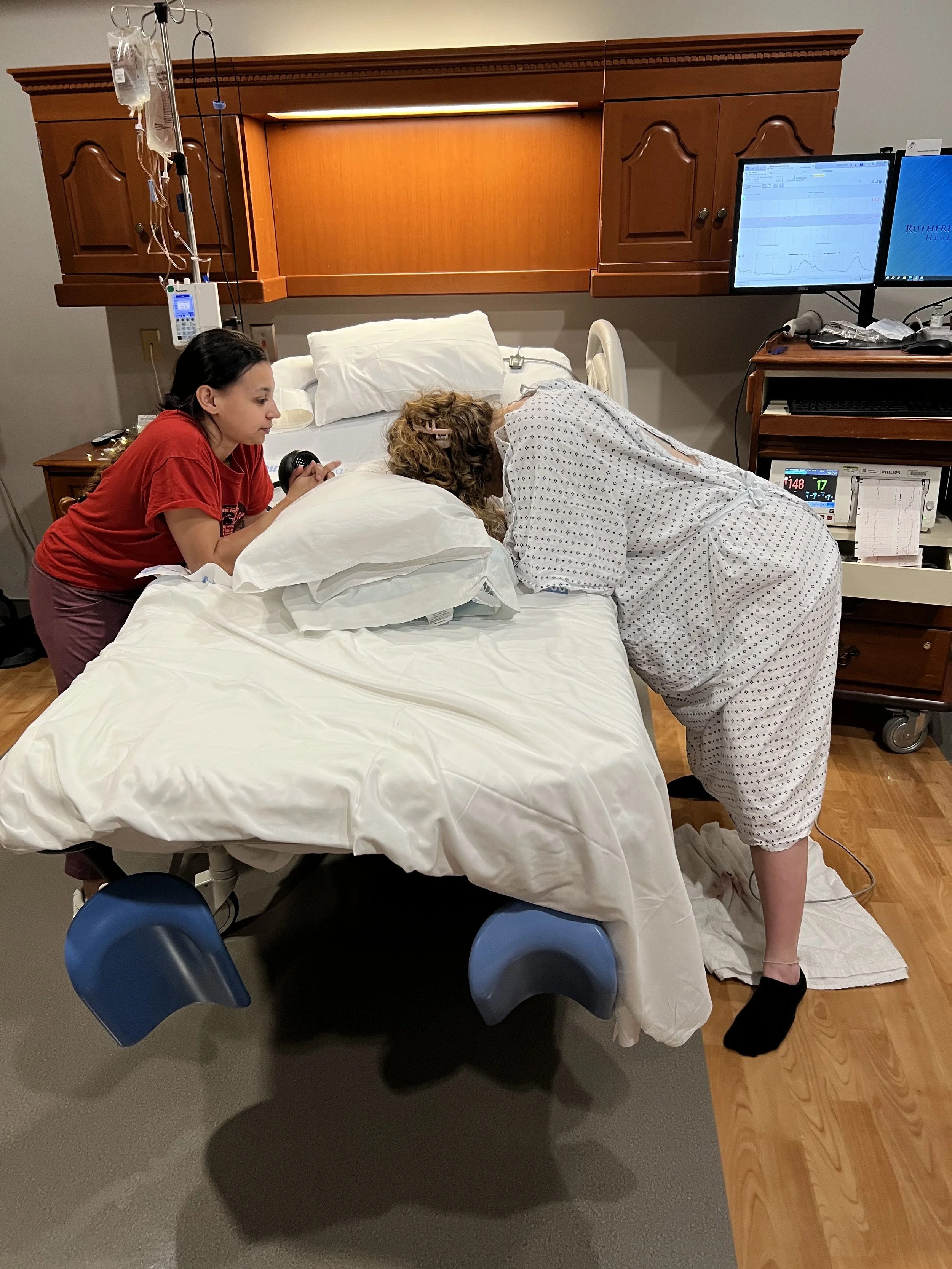 A woman in patient gown lying on hospital bed with head turned to the side, wearing a head coil and connected to medical monitoring equipment. Another woman in casual clothing holds a handheld device near the patient, possibly preparing for a medical procedure or test.