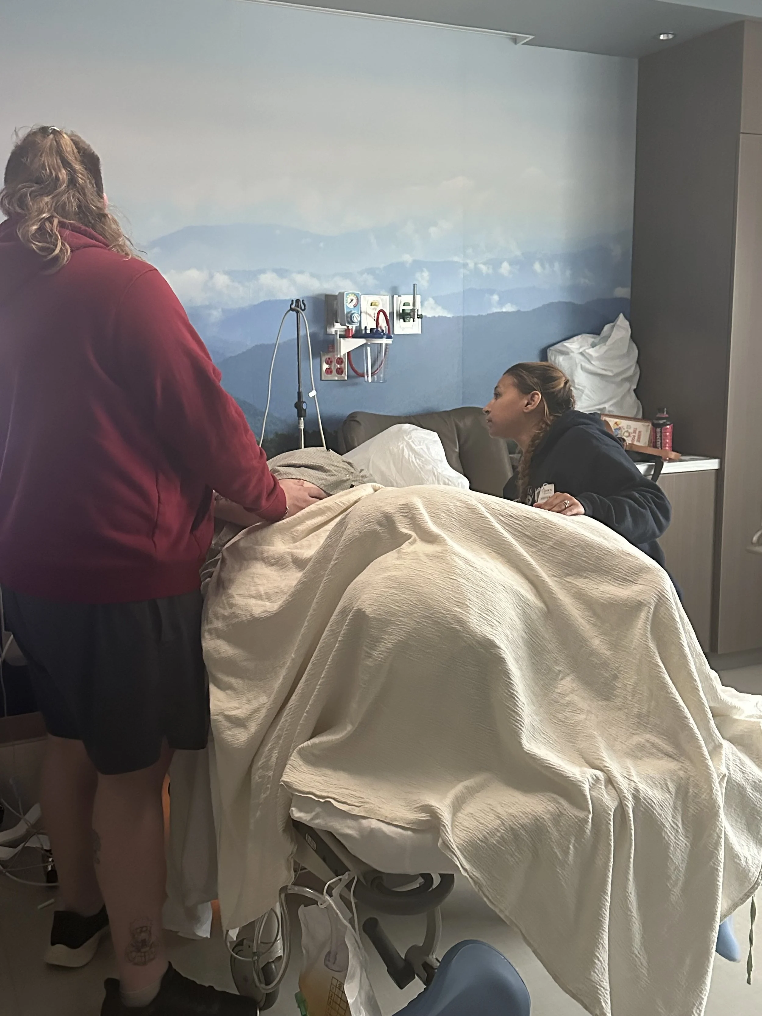 A person lying in a hospital bed with a blanket, two women standing nearby, one comforting the patient and the other sitting on a chair, with medical equipment on the wall behind