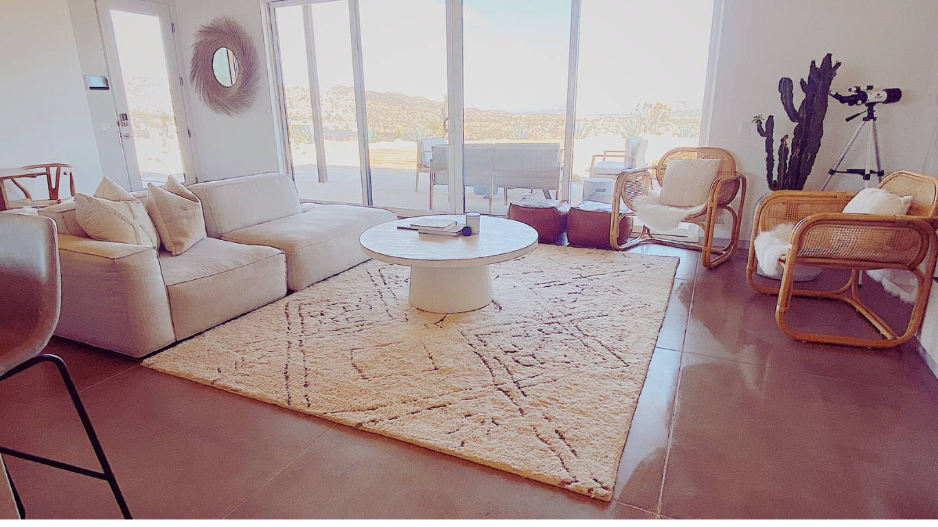Living room with a beige sectional sofa, round white coffee table, wicker chairs, and a large sliding glass door with a view of an outdoor patio and desert landscape.