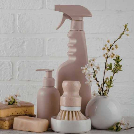 Cleaning supplies including spray bottle, pump bottle, brush, soap, and a vase with blossoms on a surface against a white brick wall