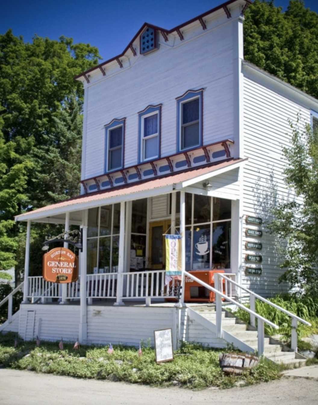 A two-story white building serving as a general store with a porch, sign, and plants in front, surrounded by green trees.