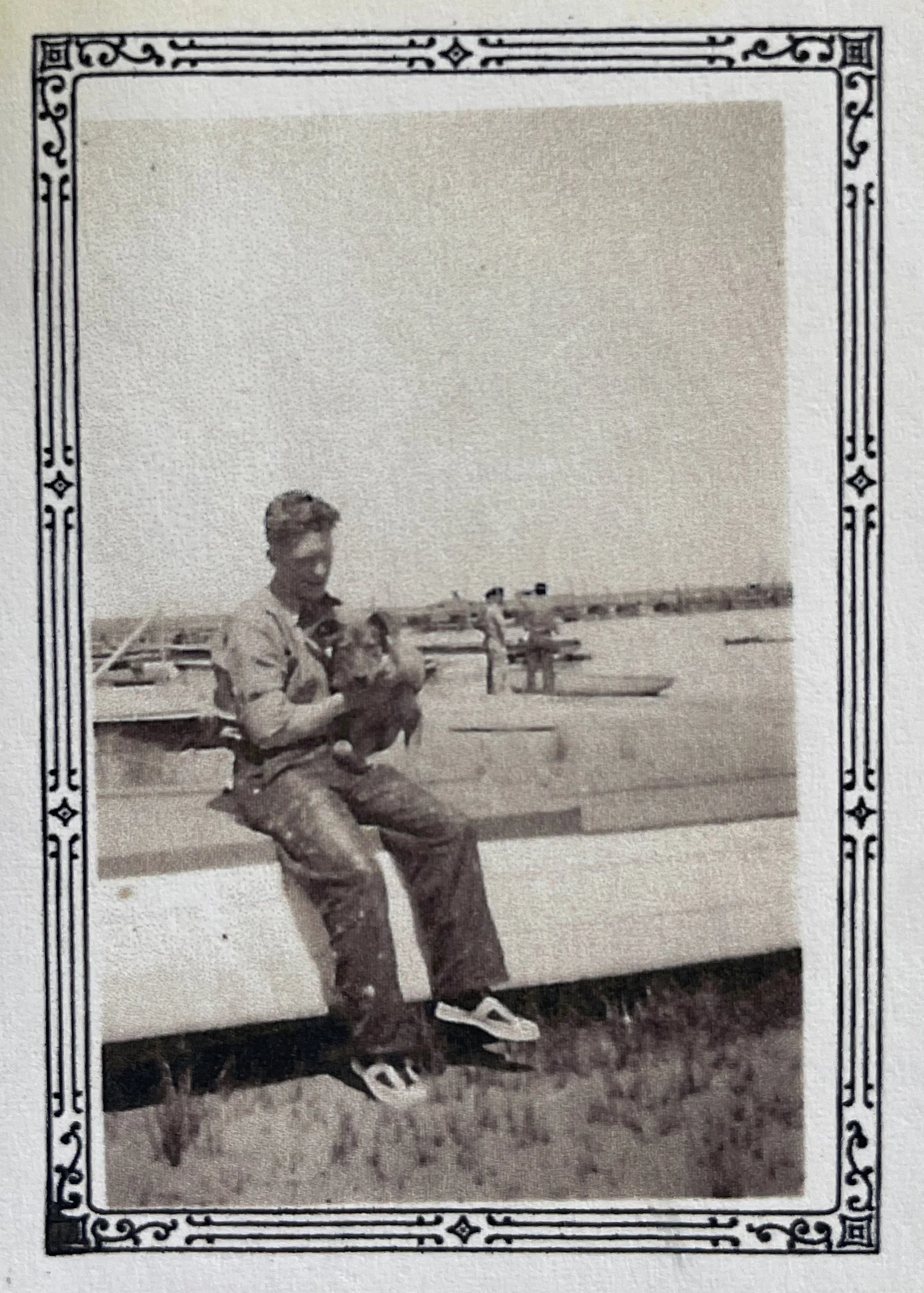 Black and white photo of a young person in a military uniform sitting on a park bench, with a field and some people in the background.