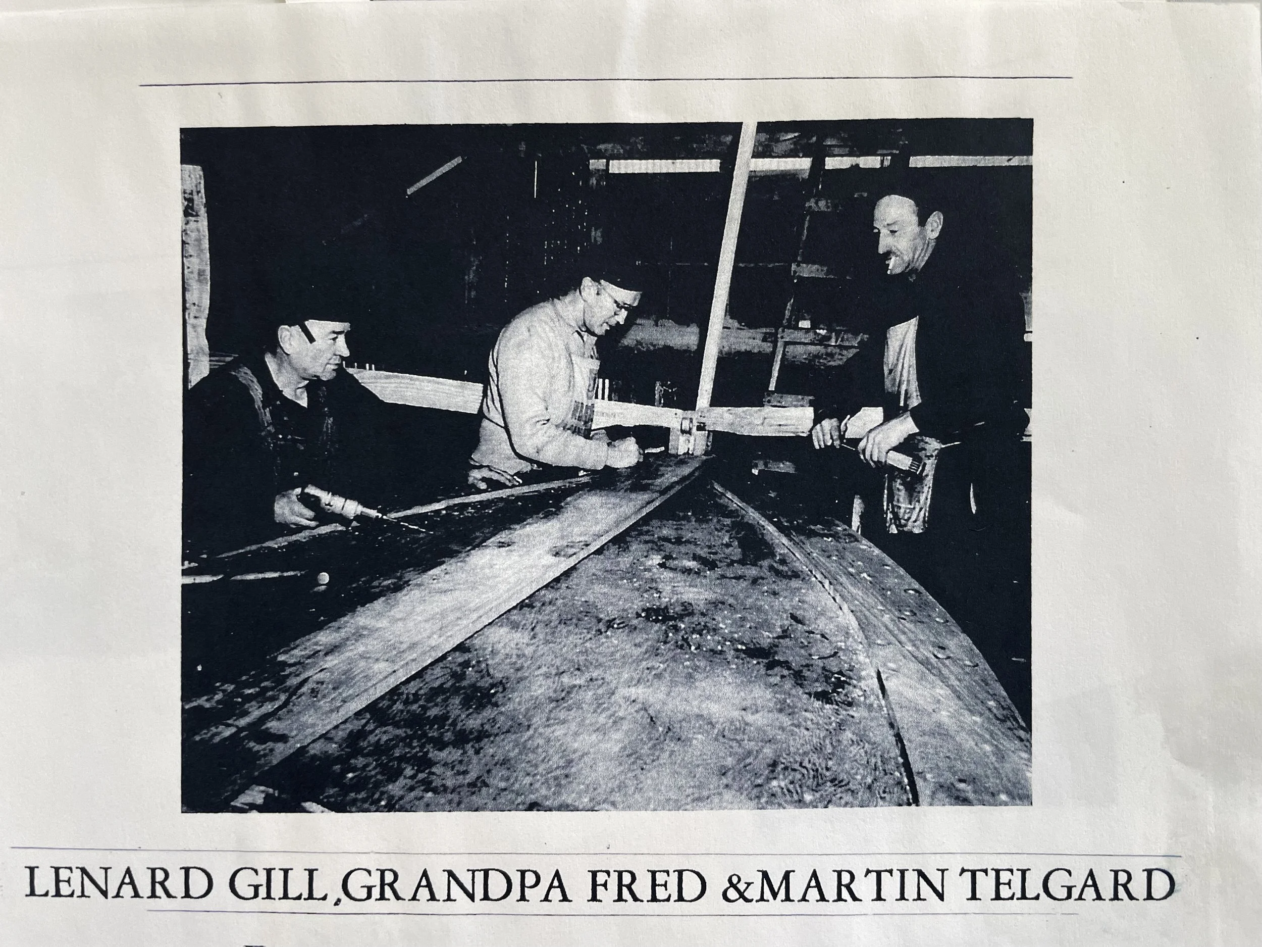 A black and white photograph of three men working on a construction or woodworking project. Two men are seated, and one man is standing, all focused on a long wood piece. The men are dressed in casual attire, and the setting appears to be a workshop 