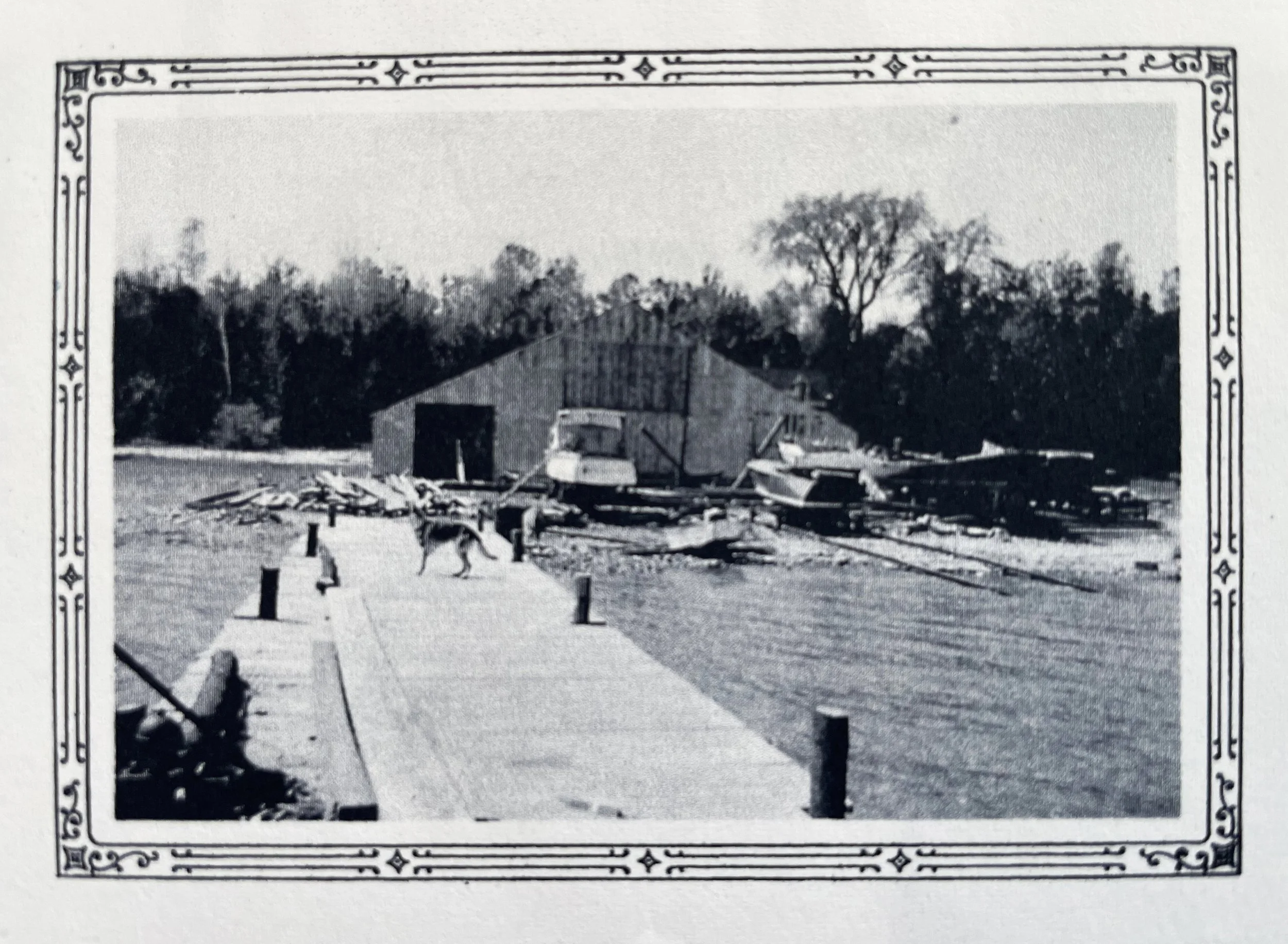A black and white photograph within a decorative border, showing a rural scene with a barn or shed, some small boats on a dock, and trees in the background.