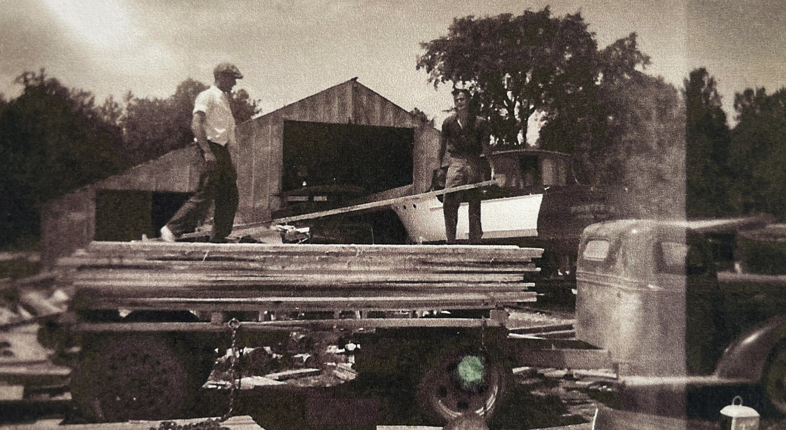 Two men standing on a flatbed truck loaded with wooden planks, with a wooden building and trees in the background, black and white photo.