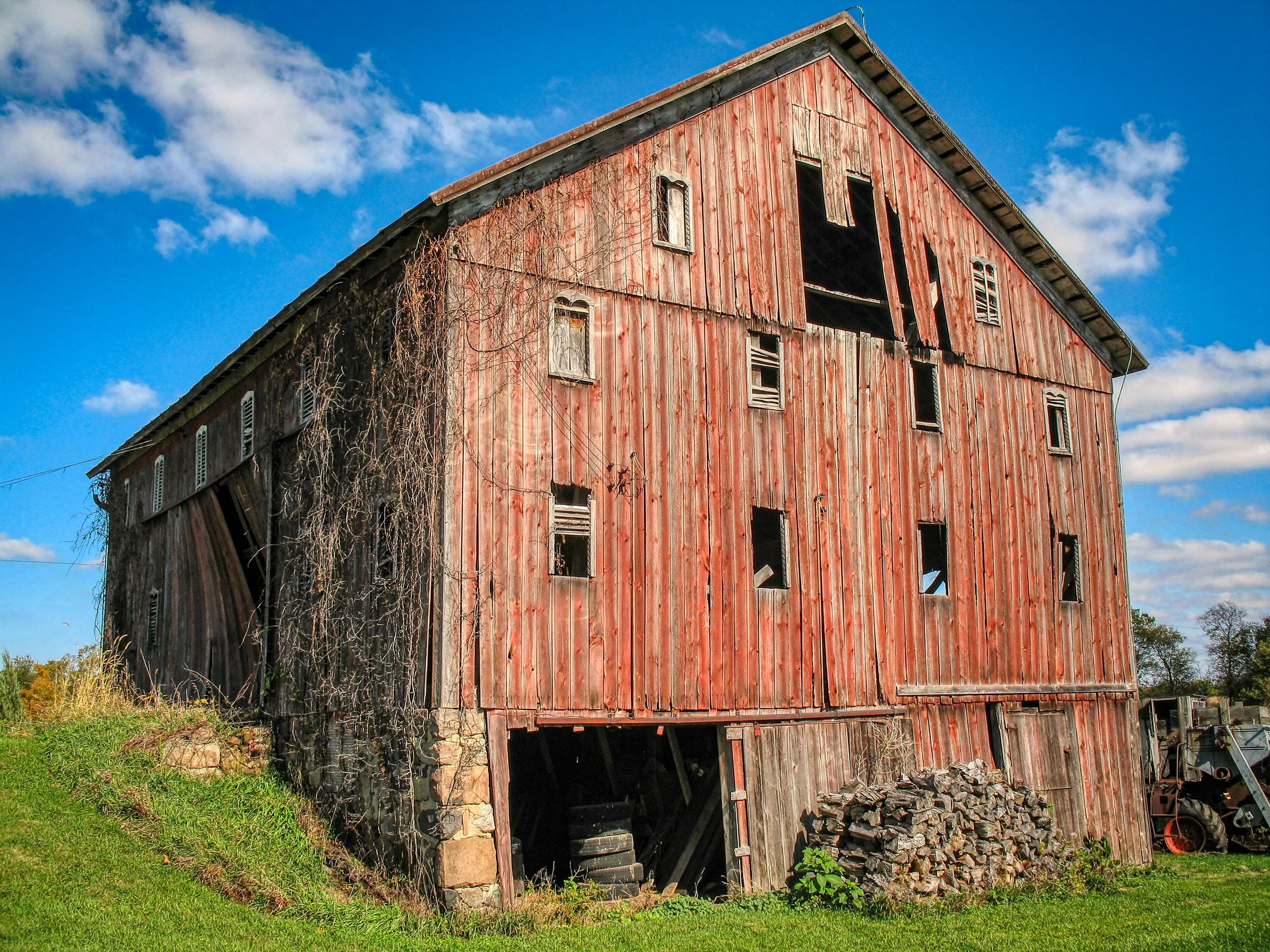 An old, weathered red barn with broken and missing window shutters stands under a blue sky with scattered clouds. The barn's lower section shows a gaping opening with a pile of stones and debris underneath. Overgrown vines and grass surround its base.
