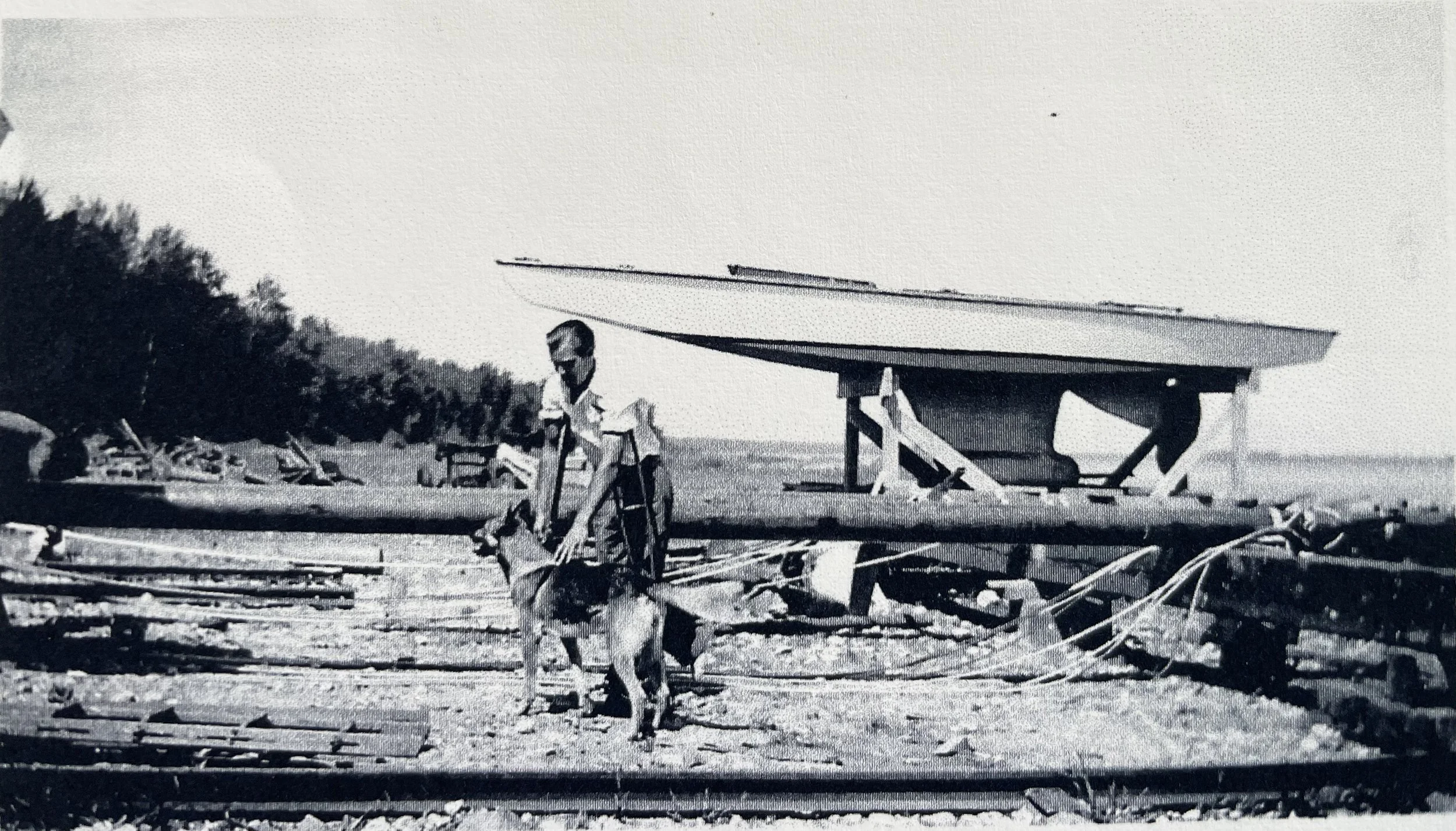 Black and white photo of a man standing outdoors with a boat on a wooden stand in the background and trees in the distance.