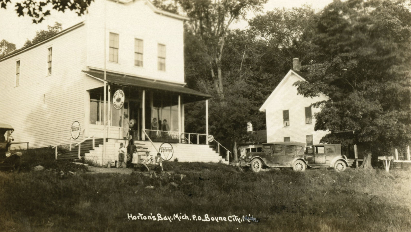 A vintage black-and-white photograph of a two-story building with a porch and a sign reading 'Horton’s Bar, Mich., P.O. Bown City.' People are standing on the porch and around the stairs, and vintage cars are parked on the grasslot in front. There are trees surrounding the buildings.
