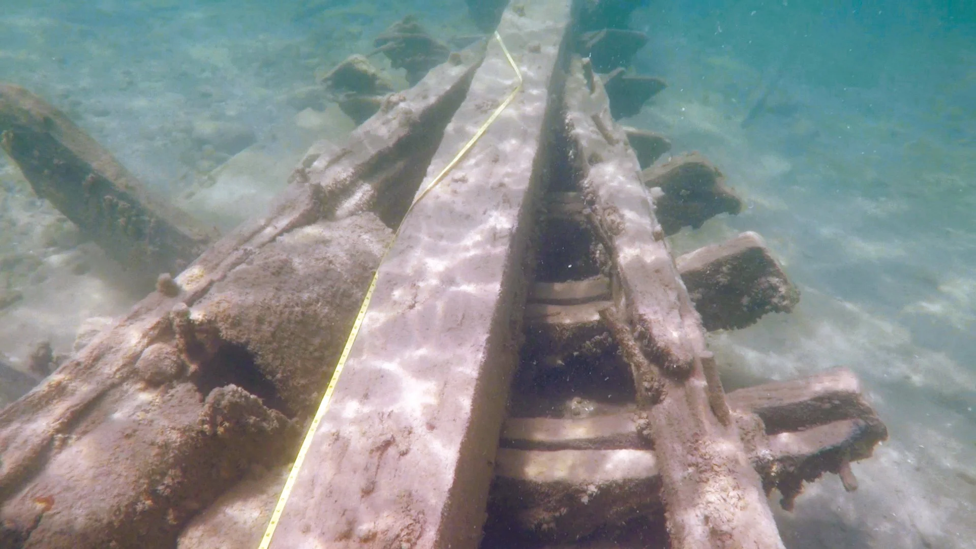 A submerged, decayed wooden structure, possibly part of a shipwreck, lying underwater in shallow clear water with a measuring tape running along its length.