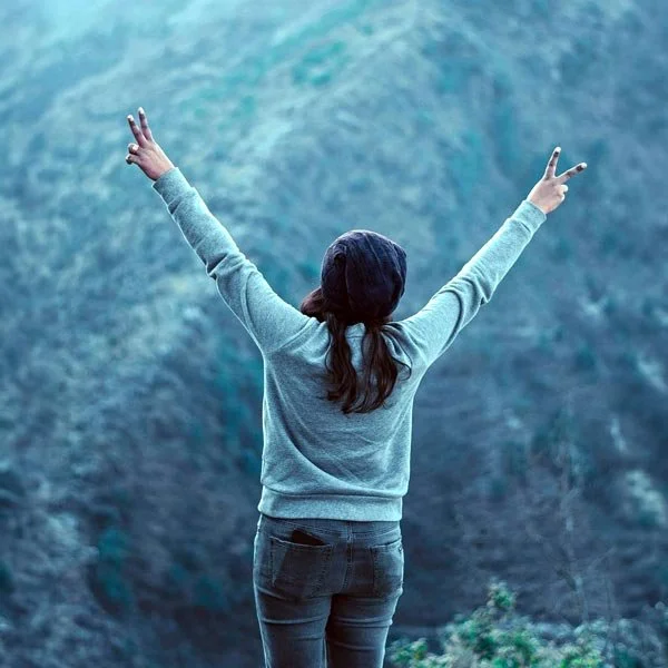 A person wearing a blue cap and gray sweatshirt making peace signs with both hands, raised in victory, standing with back to us and arms raised in front of a scenic mountain view.