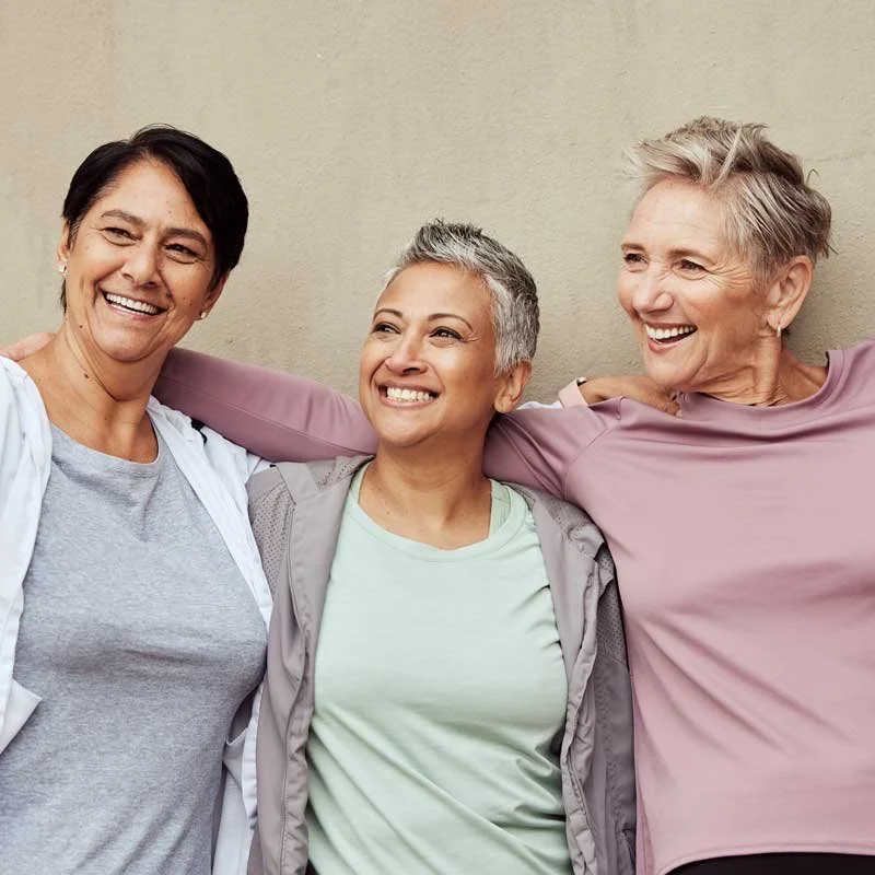 Three women smiling and standing close together with their arms around each other against a plain beige wall.