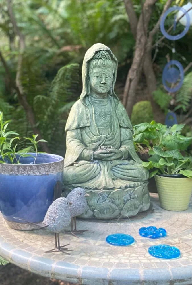 Stone garden sculpture of Kwan Yin wearing a hooded cloak, surrounded by potted plants and blue decorative stones on a patterned stone table.