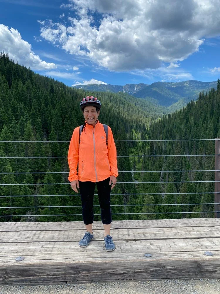 Woman smiling and wearing a helmet, orange jacket, black pants, and sneakers, standing on a wooden platform with a forested mountain landscape and blue sky with clouds in the background.