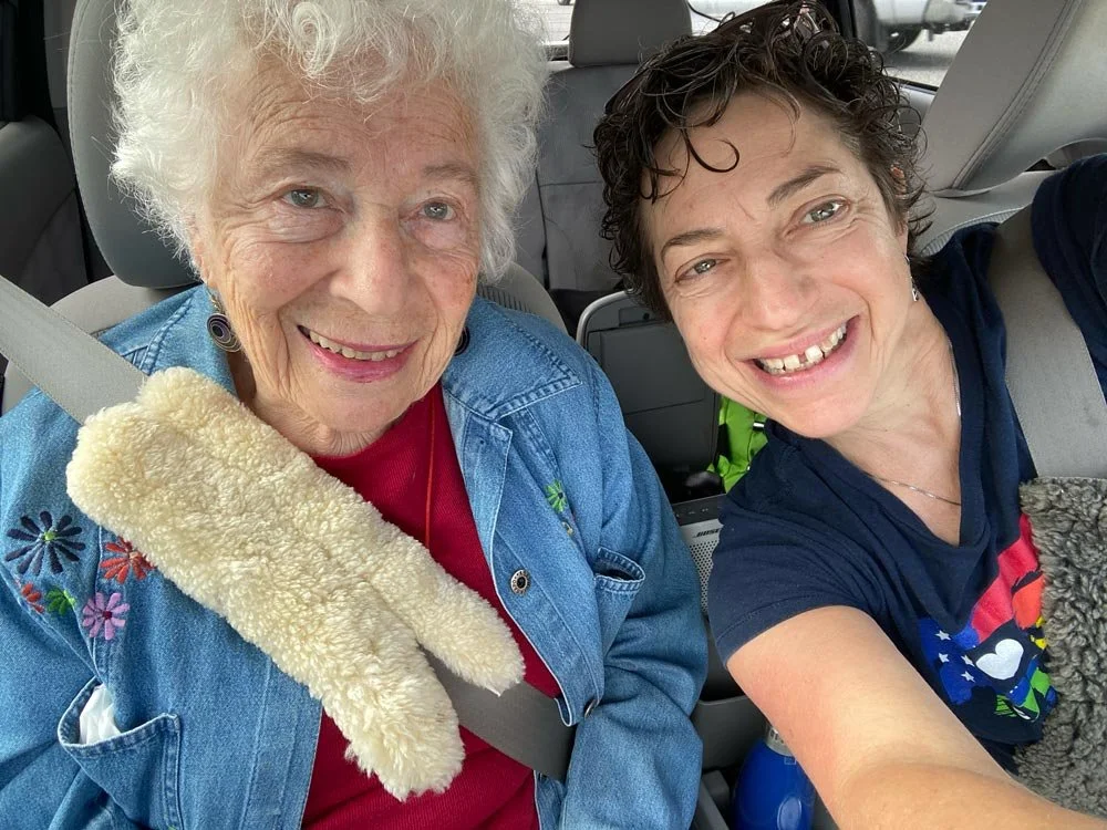 A smiling elderly woman with curly white hair and a younger woman with short curly dark hair sitting in a car. The elderly woman wears a denim jacket and a red shirt, and the younger woman is taking a selfie.