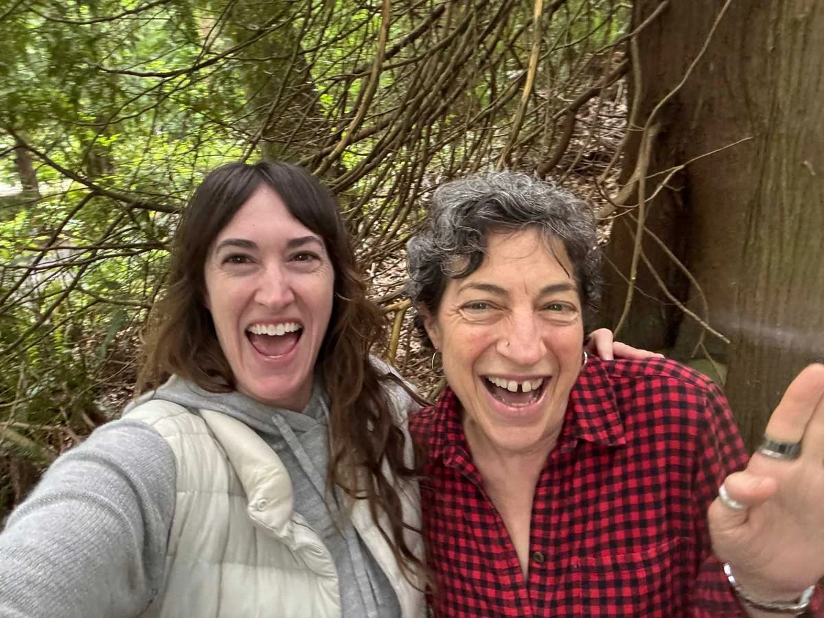 Two women smiling and posing for a selfie in a wooded outdoor area with tree branches and leaves in the background.
