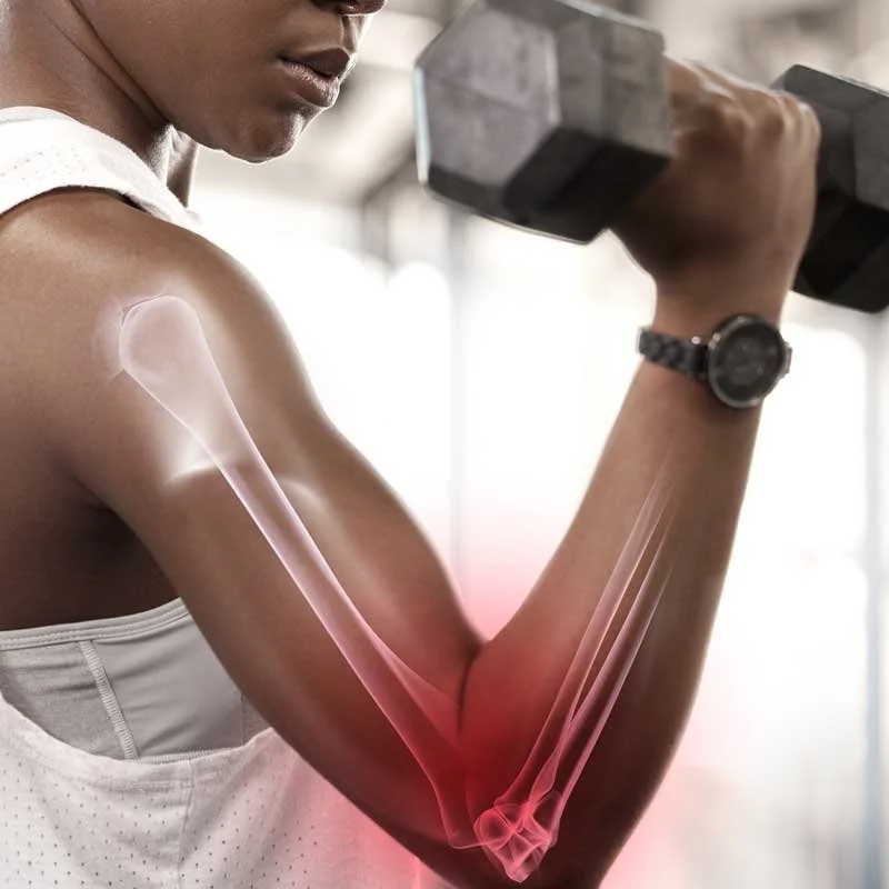 A woman lifting a dumbbell in a gym, with a digital overlay showing her elbow joint and surrounding bones in red, indicating pain or injury.