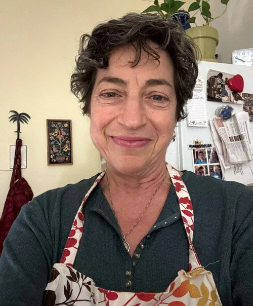 Dr. Annette Sacksteder with short, curly hair taking a selfie in a kitchen, wearing a colorful apron and dark shirt, with a smile.