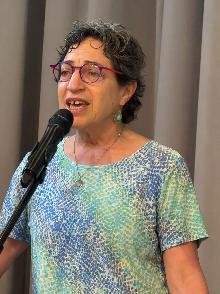 A woman speaking into a microphone at an indoor event, wearing glasses, turquoise earrings, and a colorful patterned top.