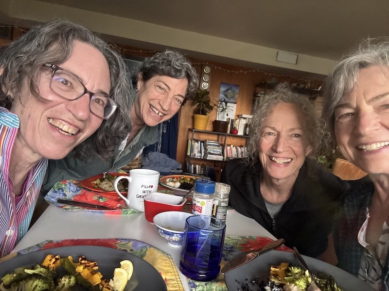 Four women smiling and sitting around a table with food, drinks, and dinnerware in a cozy indoor setting.