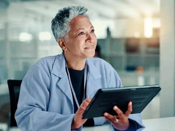 A woman with short gray hair and medium-toned skin and in a light blue lab coat, is sitting at a table in a modern, well-lit office or laboratory environment. She is holding a black tablet and appears to be looking thoughtfully into the distance.