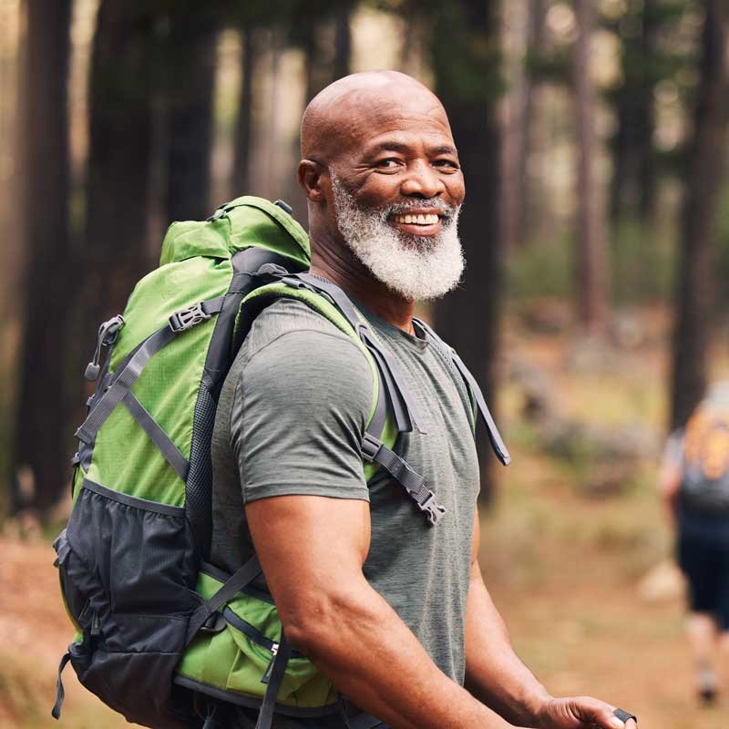 An older man with a gray beard smiling while hiking in a forest, wearing a gray shirt and a large green backpack.