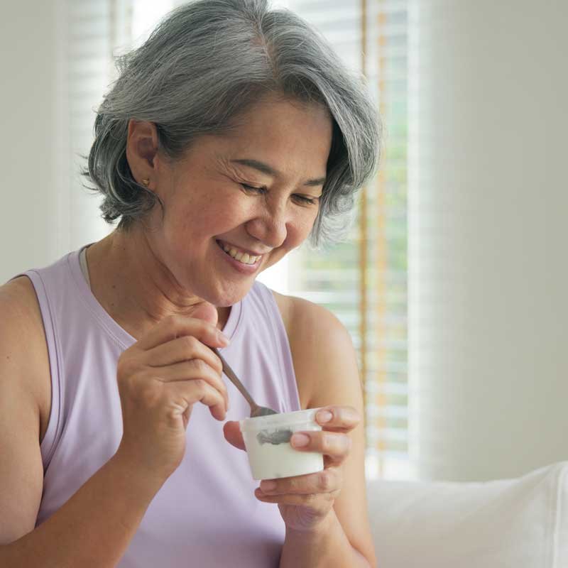 An older woman with short gray hair smiling while holding a spoon and a small cup, sitting near a window.