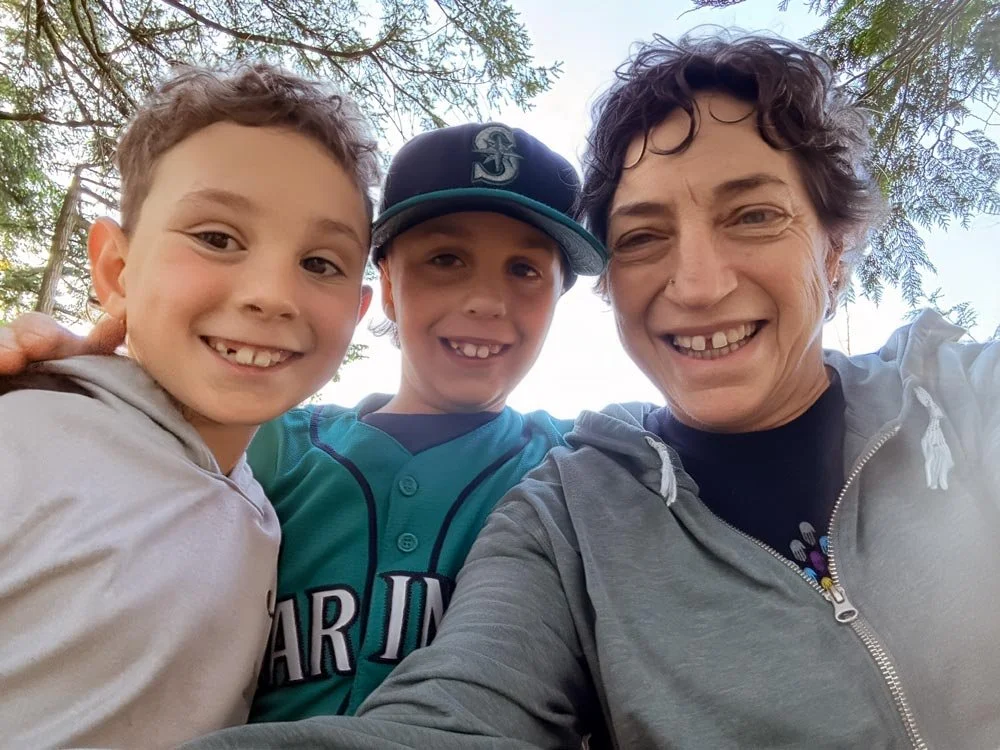 A woman and two kids smiling for a selfie outdoors under trees.