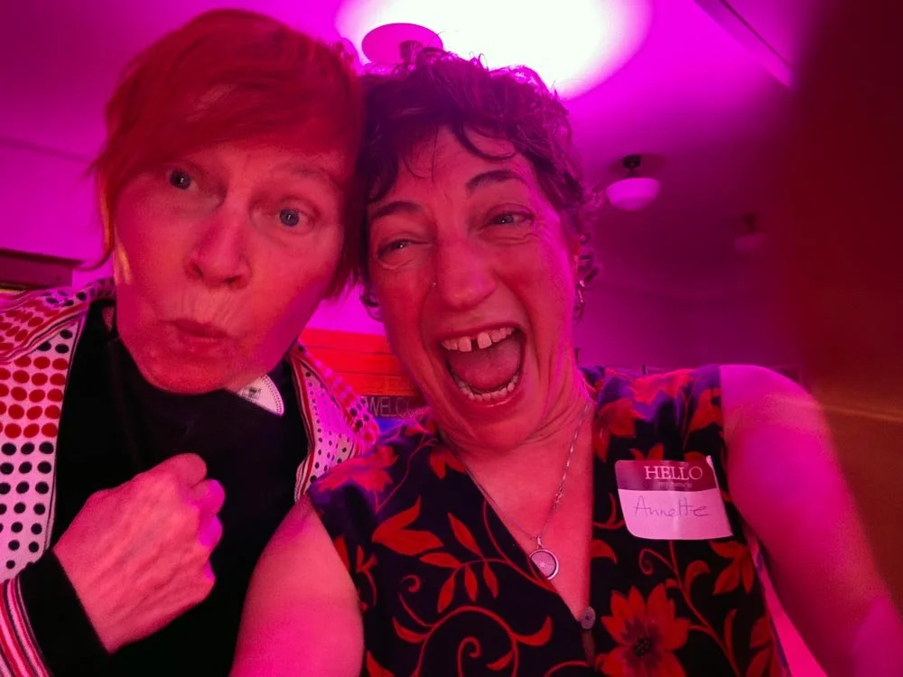 Two women taking a selfie at a party with pink lighting, one woman is wearing a shirt with floral patterns and a name tag that says "Hello, Annette," and the other woman has red hair and is making a surprised face.