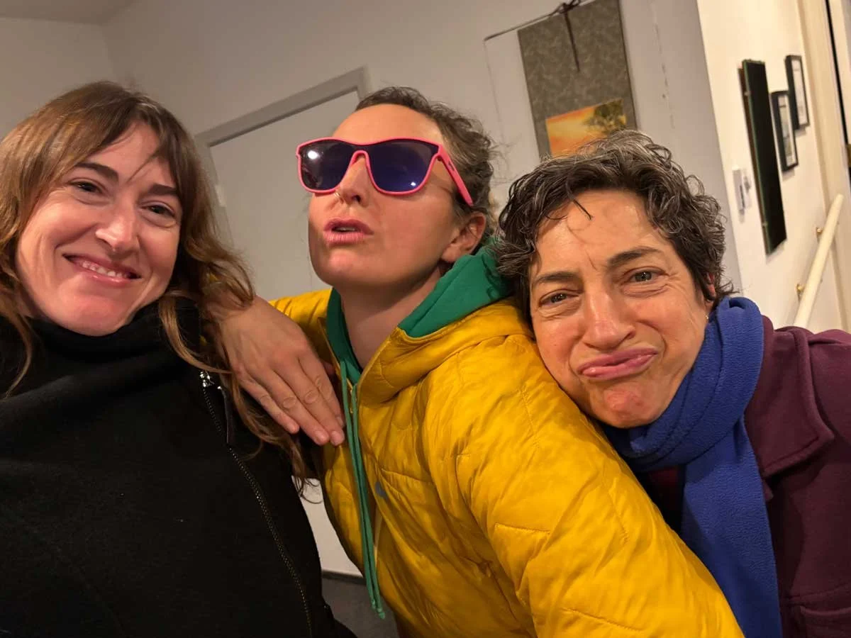 Three women taking a selfie together, one wearing pink sunglasses, another smiling, and the third making a funny face, in an indoor setting.