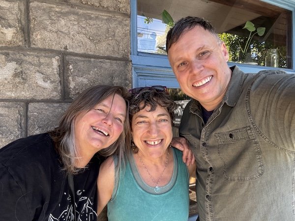 Three smiling people taking a selfie outside near a stone wall and window.