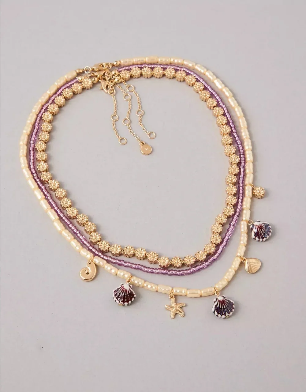 Three layered beaded necklaces with gold charms and sea shell pendants on a light background.