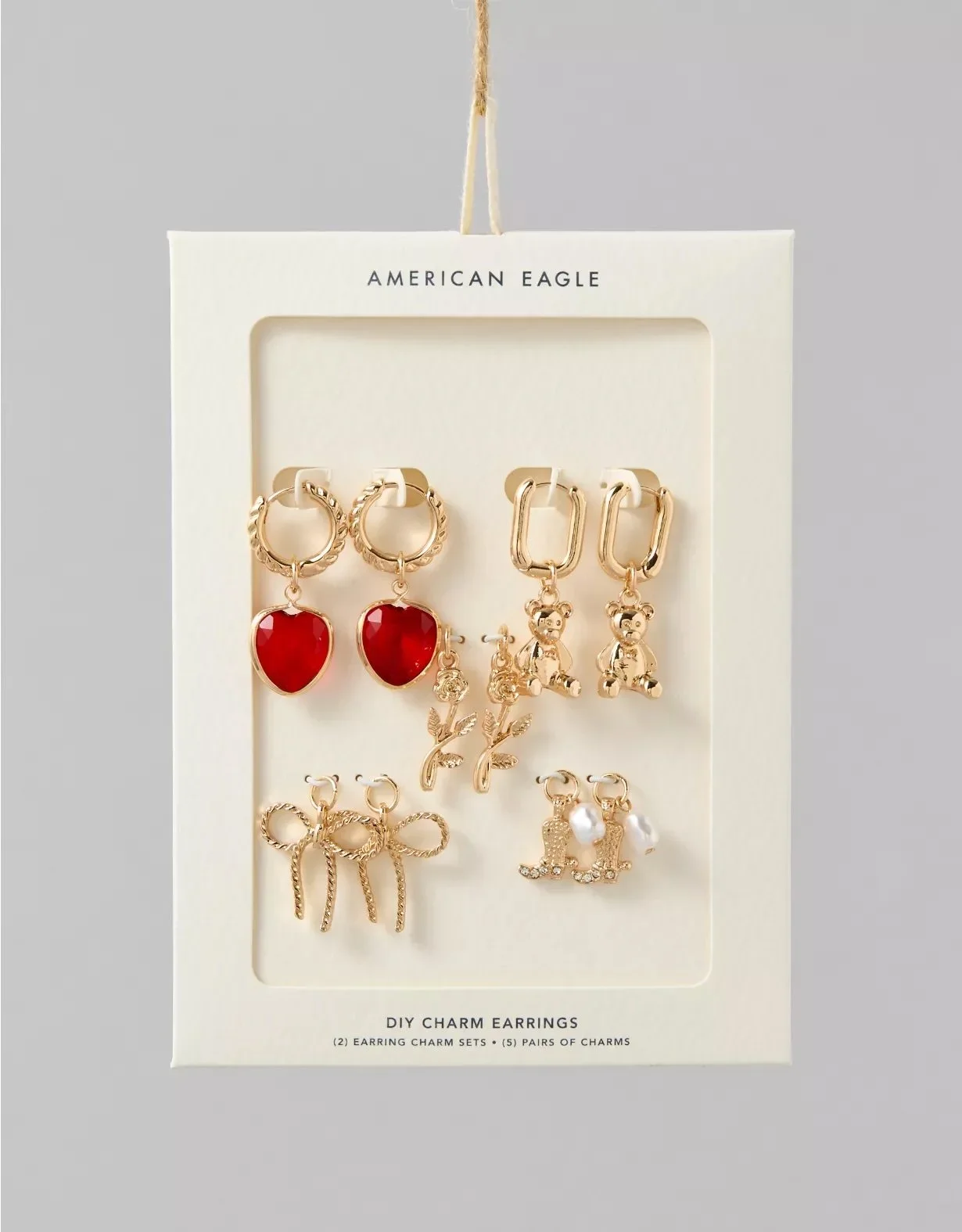 Display of five pairs of gold-colored earrings with various charms, including red hearts, teddy bears, leaves, bows, and shoes, on a white card labeled "DIY Charm Earrings" from American Eagle.