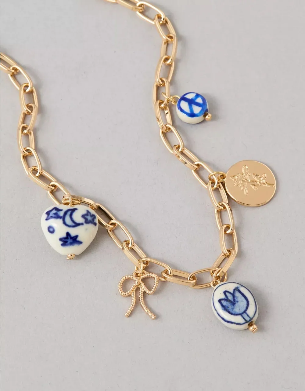 Gold chain bracelet with blue and white ceramic charms featuring celestial symbols, a bow charm, and a circular gold pendant with an engraved flower, on a light background.
