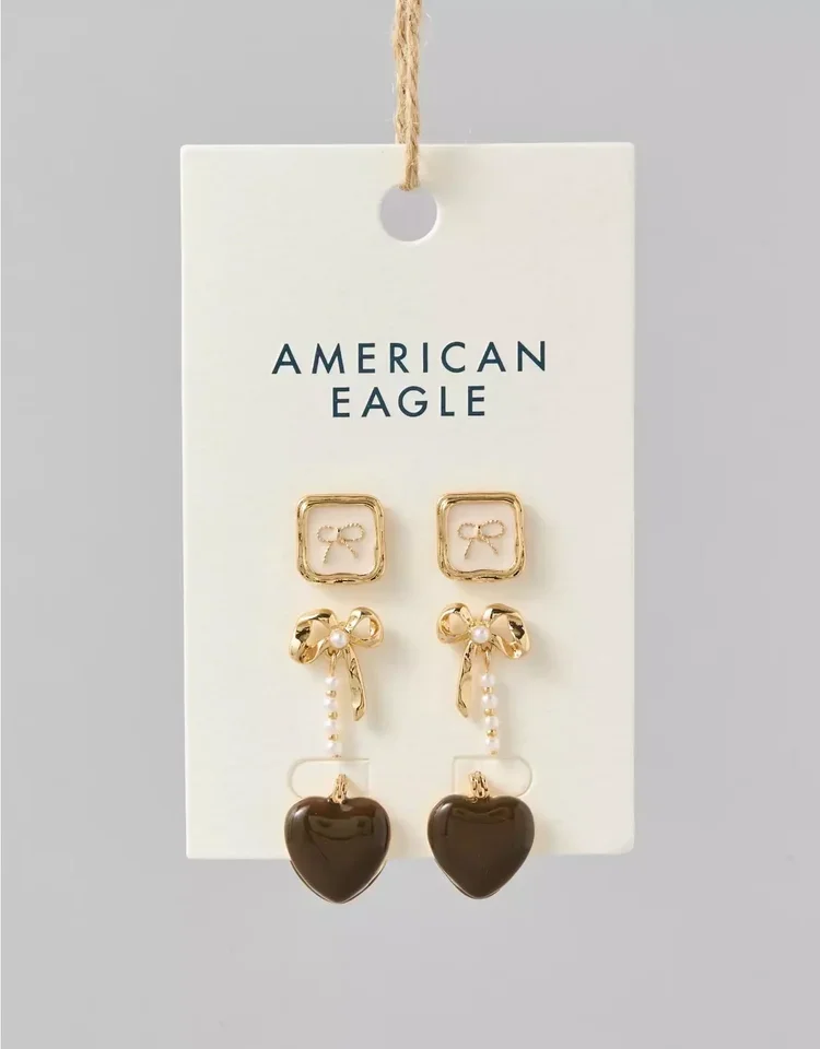 Pair of earrings with a square gold frame, a gold bow with pearl accents, and a black heart pendant, displayed on a white card with 'American Eagle' printed.