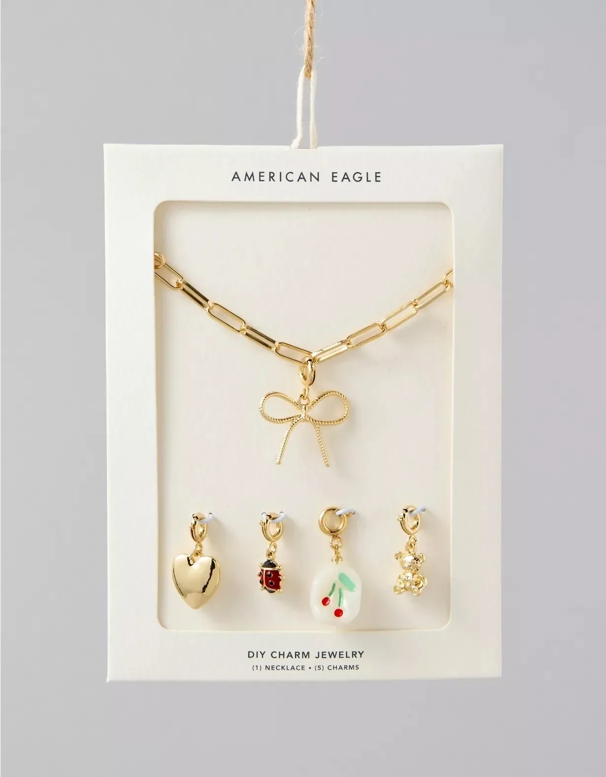 A jewelry set featuring a gold chain necklace with a bow pendant and four charm earrings including a gold heart, ladybug, cherry, and teddy bear, displayed on a white cardboard with "American Eagle" and "DIY Charm Jewelry" printed at the top and bottom.