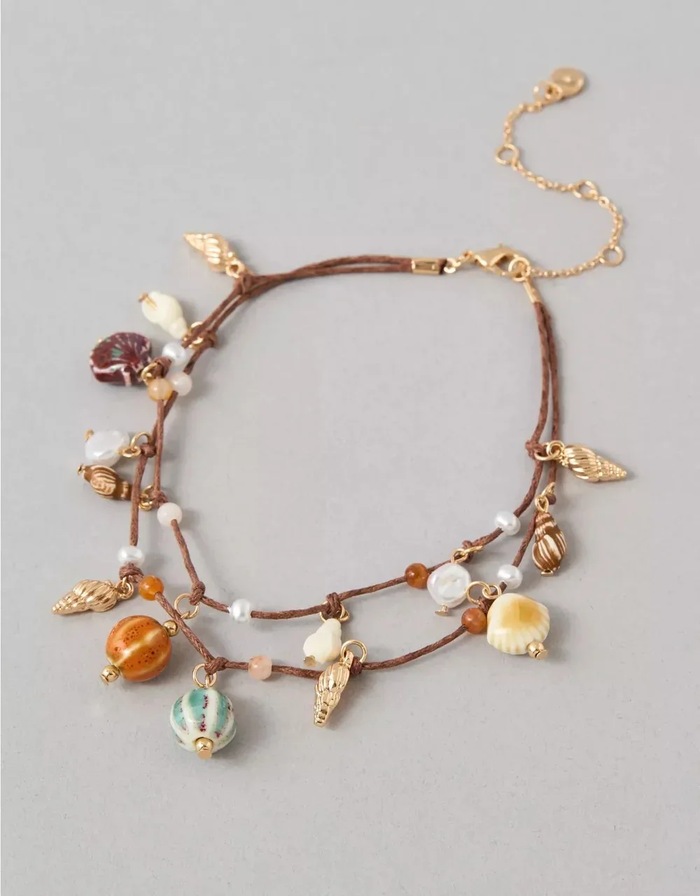 A layered necklace with shells, beads, and gold charms on brown cords, displayed on a gray background.
